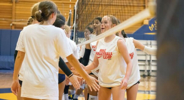 A group of young athletes wearing white shirts line up and shake hands over a volleyball net in a gymnasium, displaying good sportsmanship after a game.
