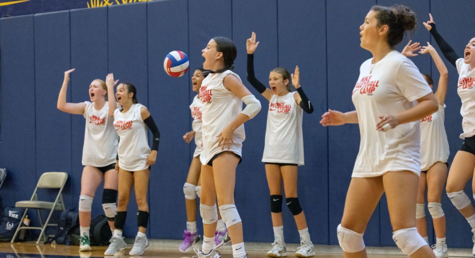 A group of girls in matching white shirts and knee pads cheer excitedly in a gym as one volleyball player stands in the center, tossing a ball to serve.