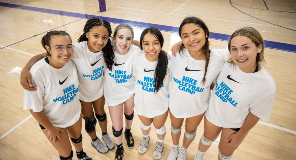 Six young women in white Nike Volleyball Camp shirts stand together in a gym, smiling and posing for the camera, with a volleyball court visible in the background.