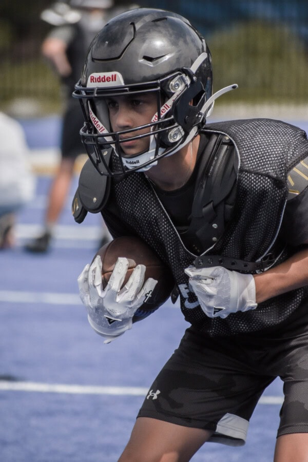 A young football player in black gear and helmet holds a football while preparing to run during practice on a blue field. Other players are blurred in the background.