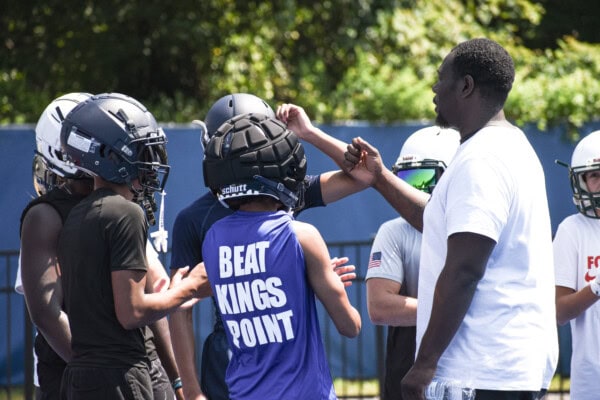 A group of young football players in helmets and athletic gear huddles together outdoors, bumping fists with a coach in a white shirt, as trees and a blue fence are visible in the background.