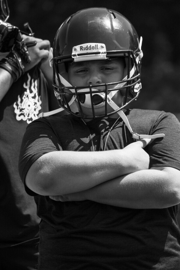 A young boy wearing a football helmet and t-shirt stands with his arms crossed, looking confidently at the camera. Another person in sports gear is partially visible beside him. The image is in black and white.